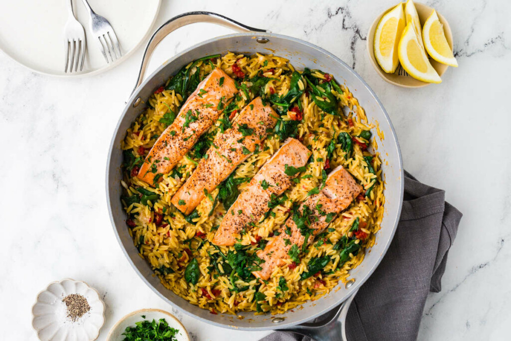 A one pan salmon and orzo recipe in a skillet next to sliced lemon.