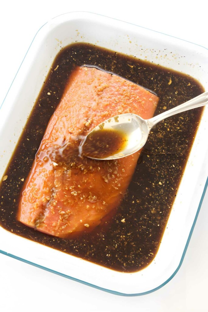 Raw salmon fillet in white dish, spooning dark marinade over fish.