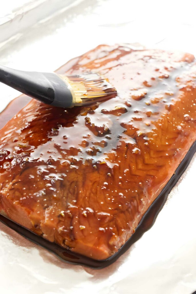Brush applying glossy brown glaze to raw salmon fillet on foil.