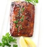 Glazed salmon fillet with parsley and lemon wedges on white plate.