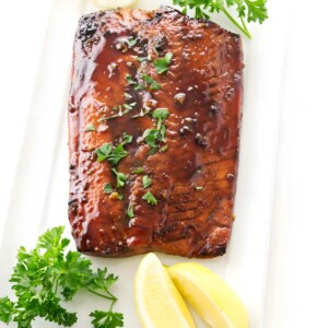Glazed salmon fillet with parsley and lemon wedges on white plate.