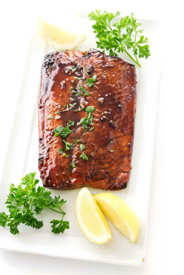 Glazed salmon fillet with parsley and lemon wedges on white plate.