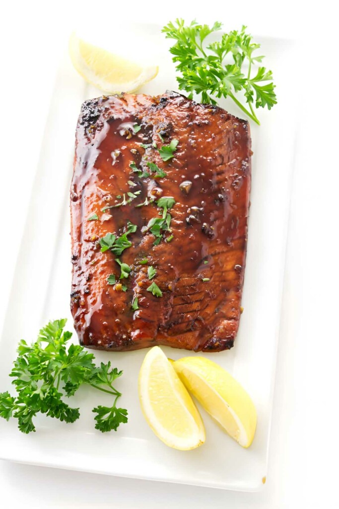 Glazed salmon fillet with parsley and lemon wedges on white plate.