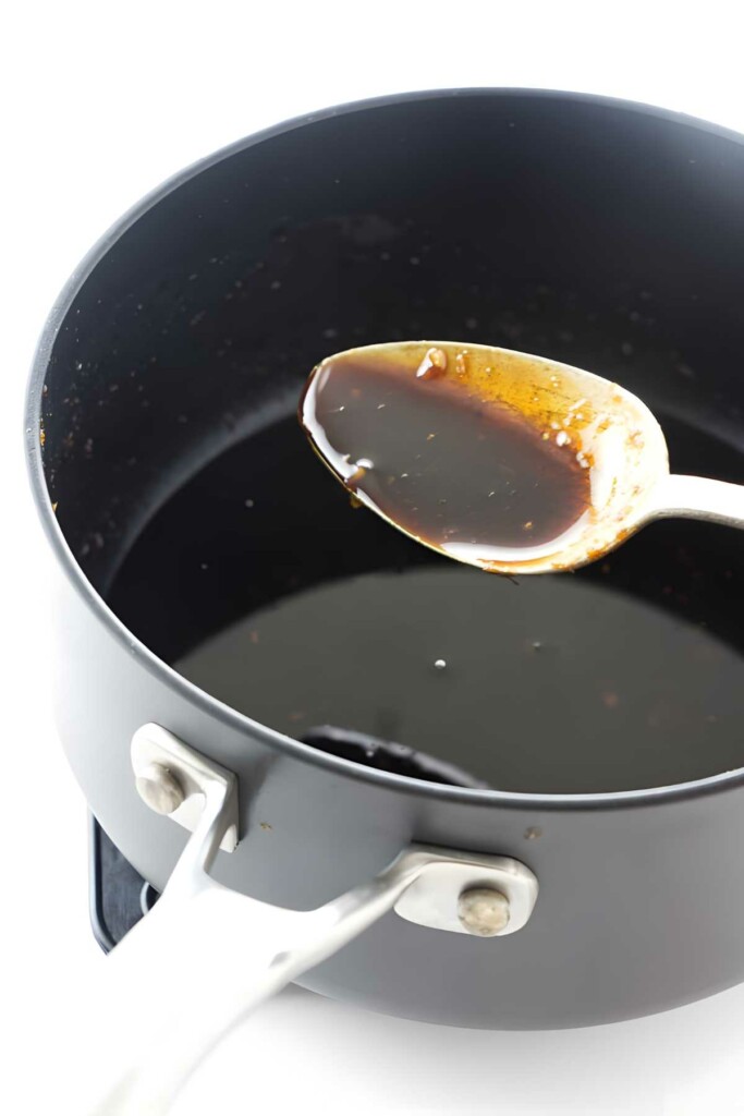 Metal spoon with dark sauce above black saucepan containing more sauce.