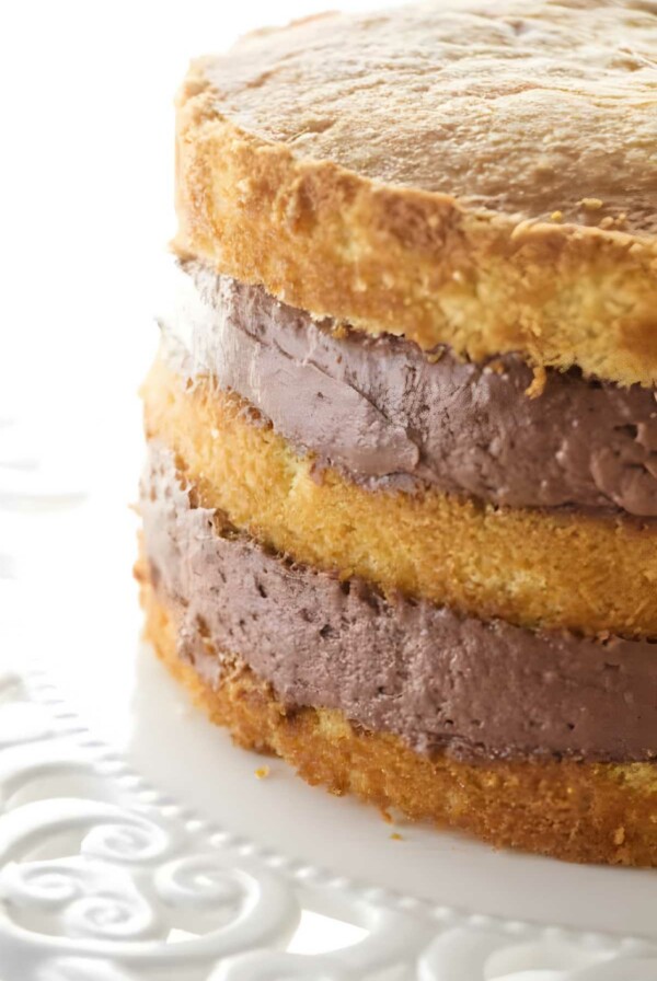 Close-up of layered vanilla sponge cake with chocolate frosting on white plate.