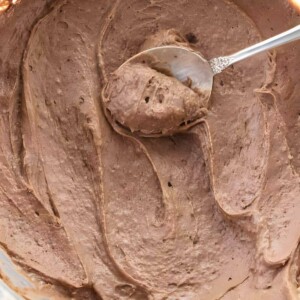 Glass bowl of creamy chocolate frosting stirred with a metal spoon.
