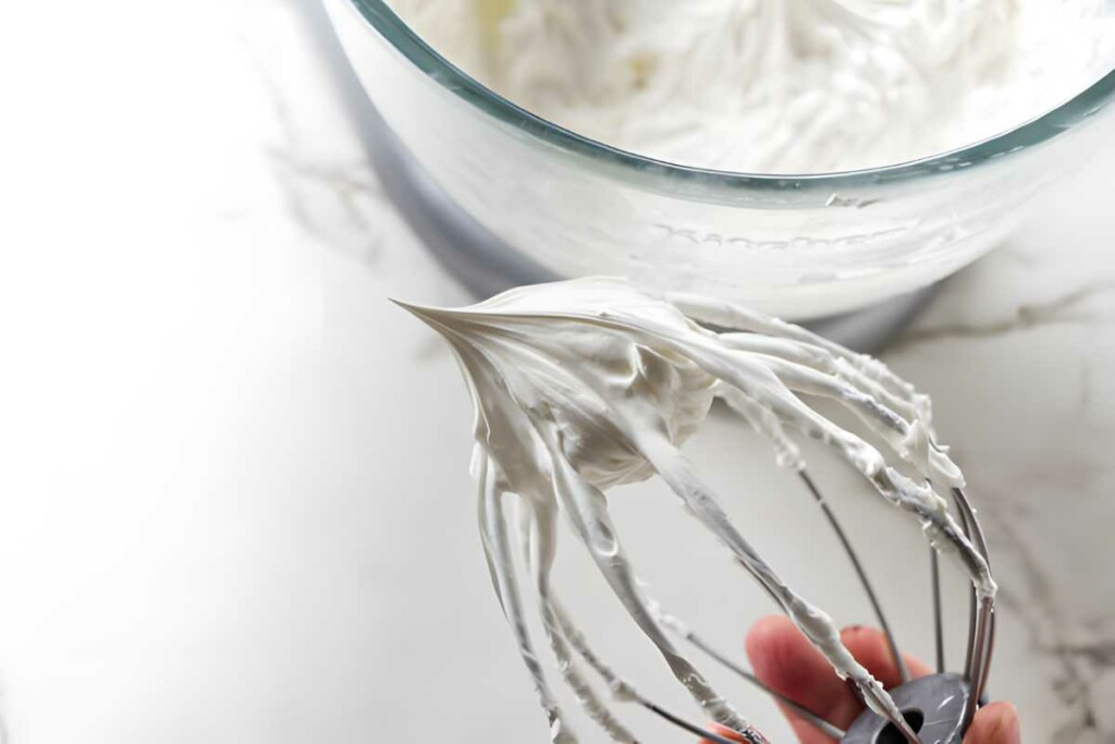 Hand holding whisk with stiff, glossy meringue; bowl of meringue behind.