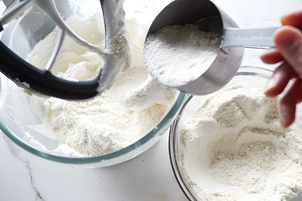 Hand pouring flour into whipped egg whites in mixer bowl on countertop.