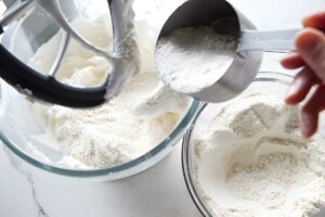 Hand pouring flour into whipped egg whites in mixer bowl on countertop.