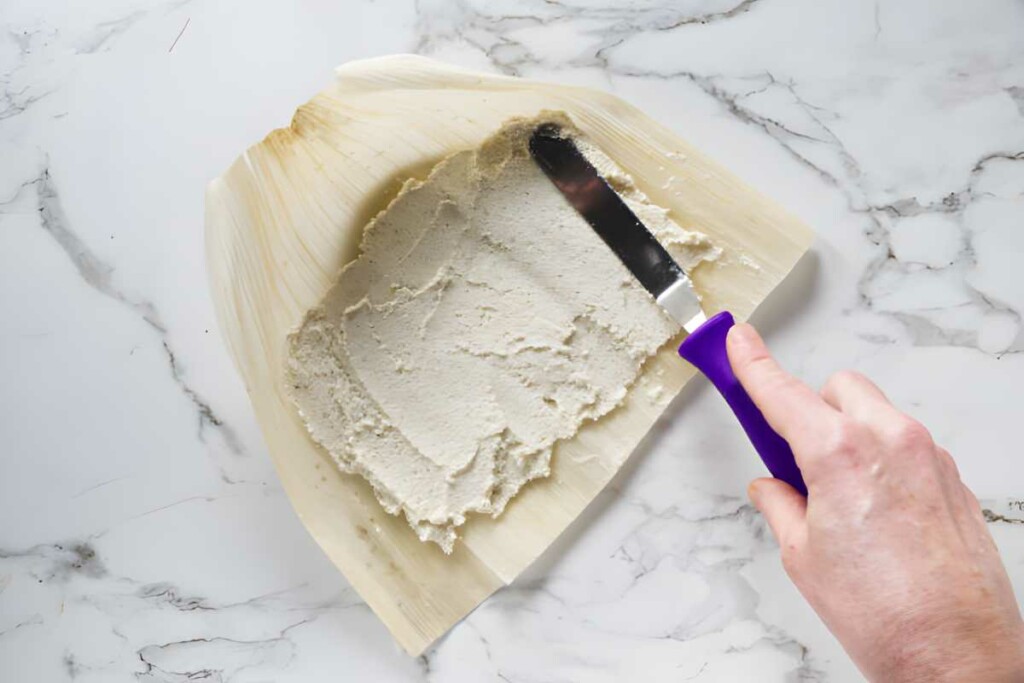 Hand spreading masa on corn husk with spatula, white marble background.