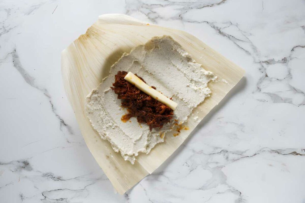 Unwrapped tamale with masa, cheese, and red meat on a corn husk.