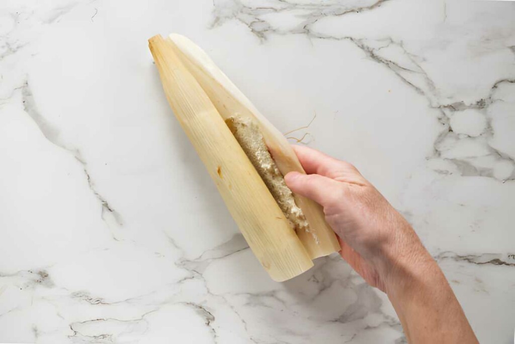 Hand holding corn husk with masa dough on marble surface.