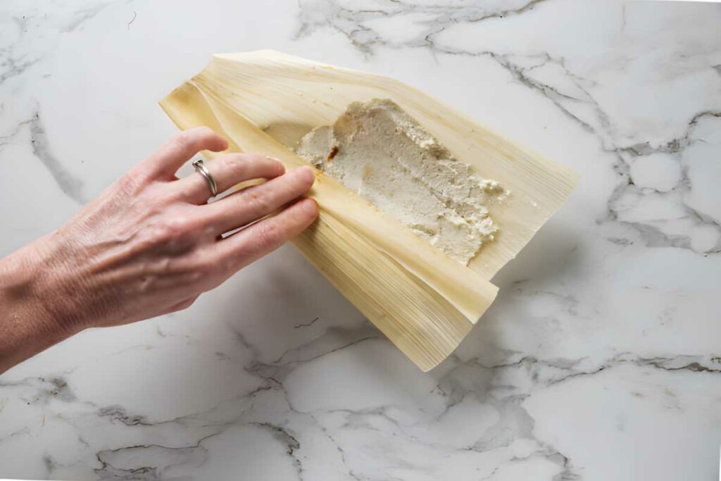 Hand spreading masa dough on corn husk for tamales on marble surface.