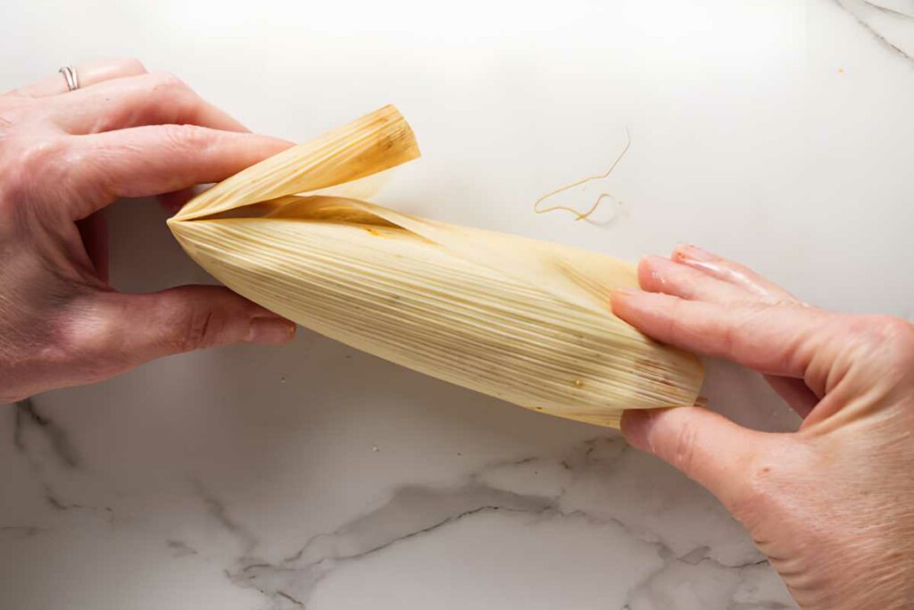 Hands folding corn husk around tamale filling on white marble surface.