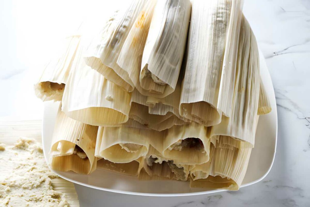 Stack of uncooked tamales in corn husks on a white plate.
