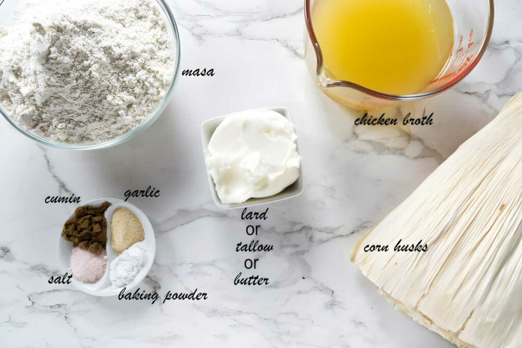 Tamale ingredients arranged on marble: masa, broth, lard, corn husks, spices.