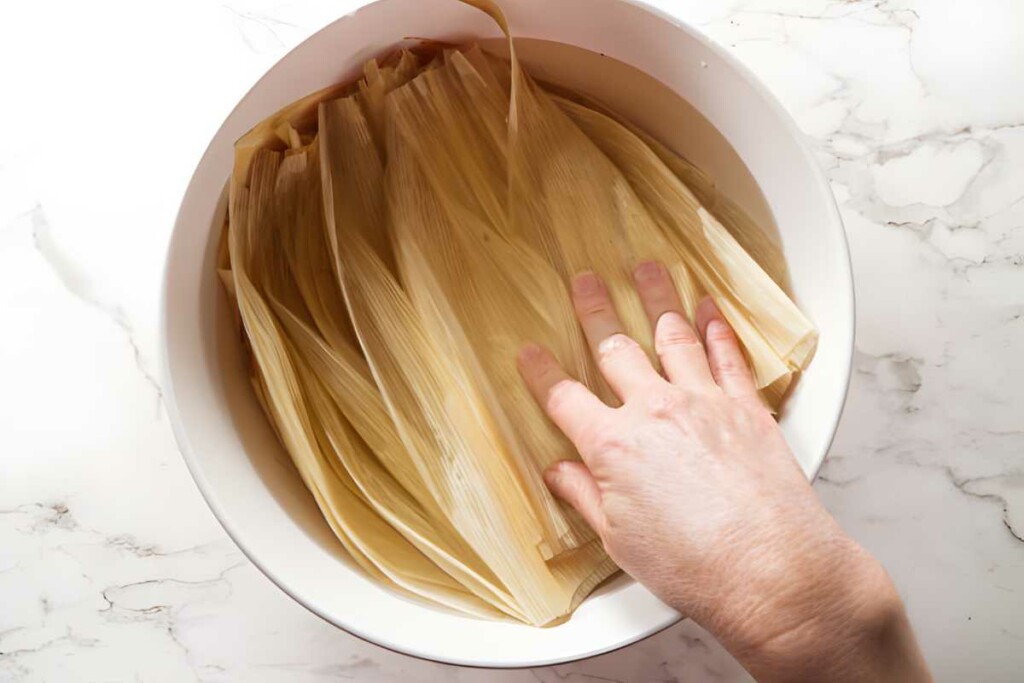 Hand pressing corn husks in water-filled white bowl on marble surface.