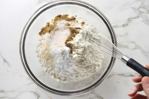 Hand whisking flour and spices in glass bowl on marble surface.