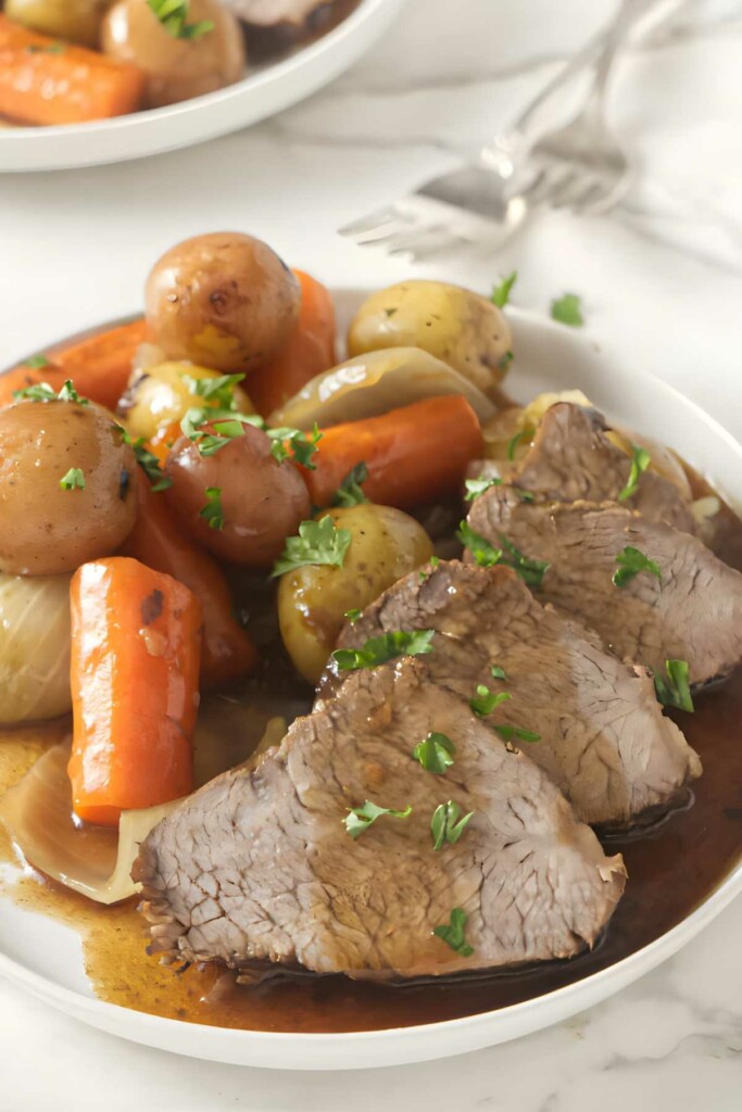 Roast beef with gravy, baby potatoes, carrots, onions, and parsley garnish.