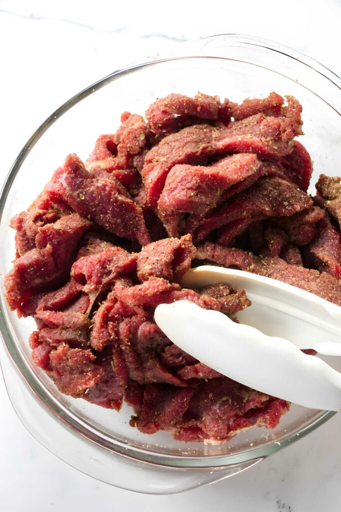 Thinly sliced seasoned beef strips held by tongs in glass bowl.
