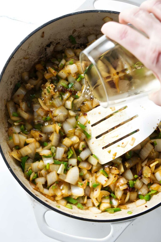 Hand pours liquid into pot of sautéed onions, peppers, and spices.