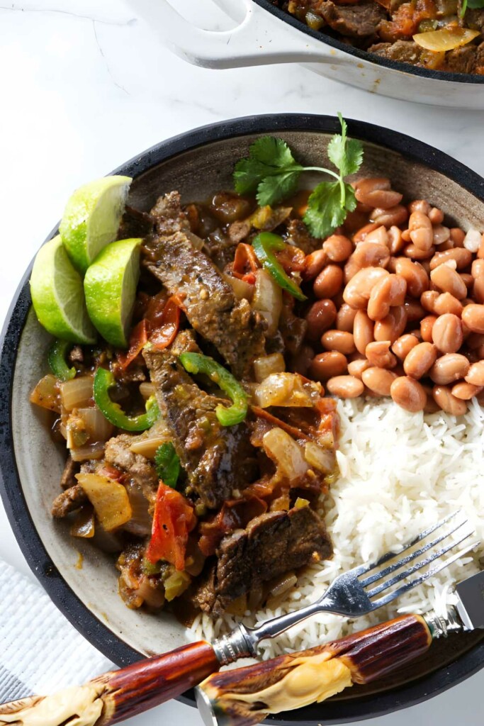 Plate of sliced beef with rice, beans, vegetables, lime, and cilantro.
