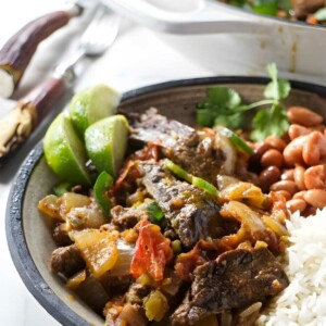 Bowl of beef stew with rice, beans, lime; pot in background.