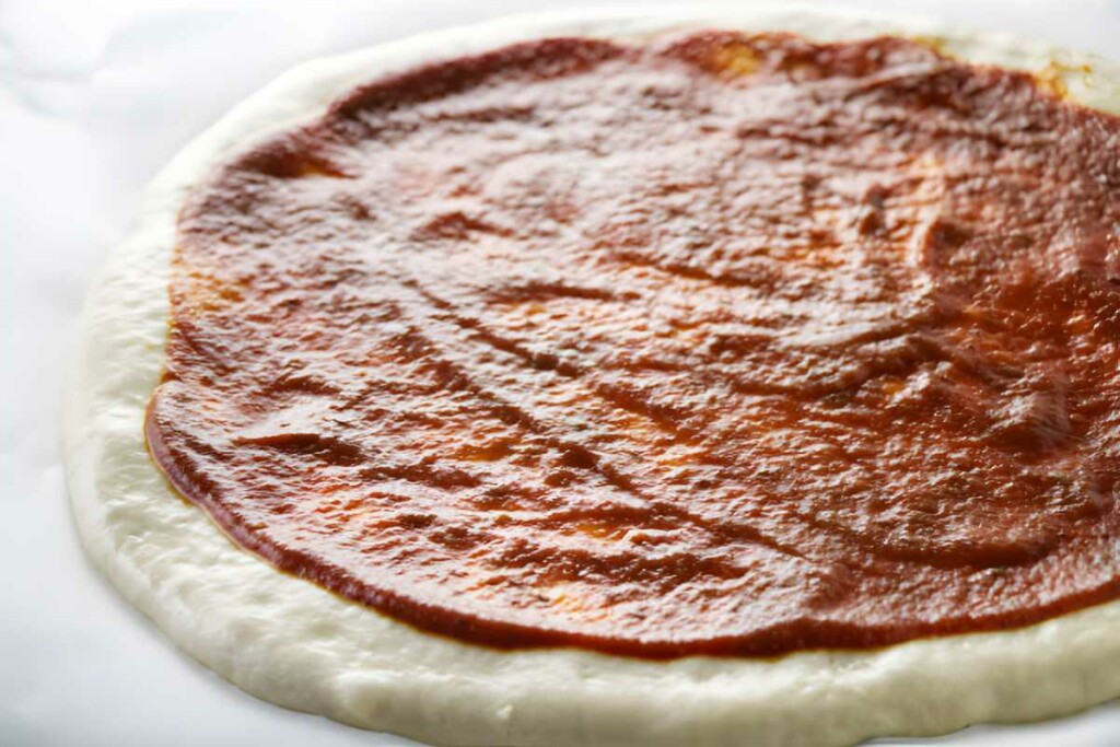 Uncooked pizza dough with tomato sauce spread, ready for toppings.