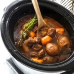 Wooden spoon stirs beef stew with vegetables in black slow cooker.