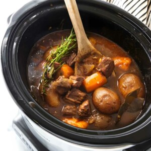 Wooden spoon stirs beef stew with vegetables in black slow cooker.