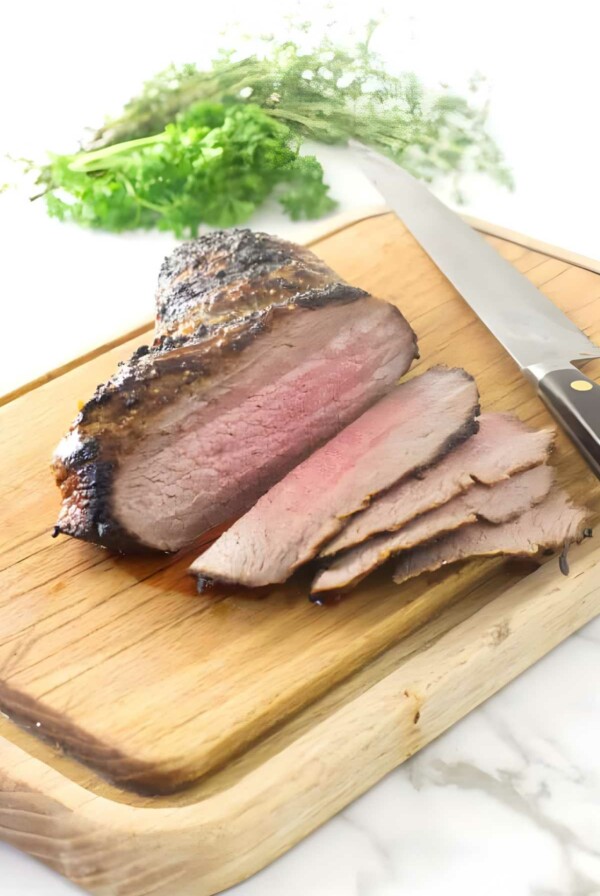 Sliced tri tip on cutting board with knife and parsley in background.