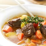 Close-up of lamb soup with vegetables, bread in background.