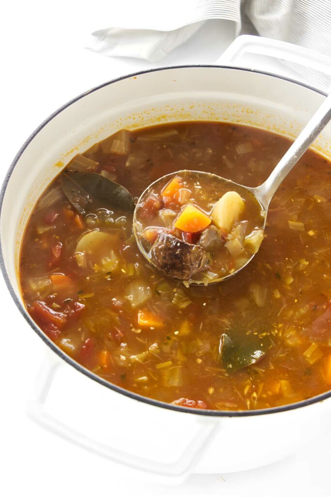 Ladle lifting chunky lamb and vegetable soup from a white pot.