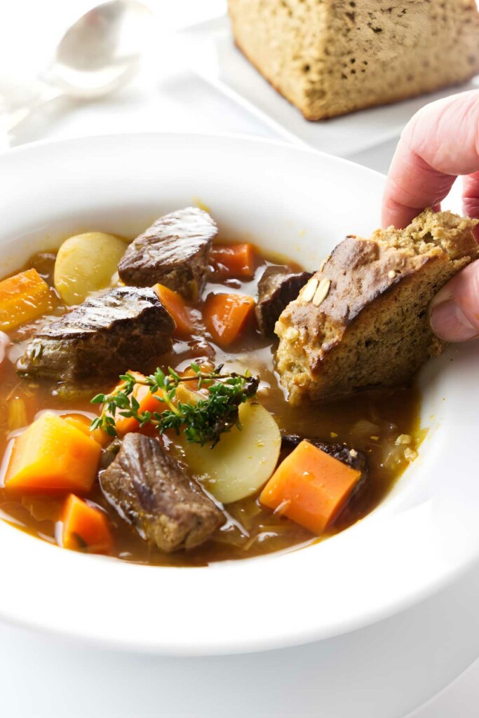 Hand dipping bread into lamb stew with vegetables; loaf of bread behind.
