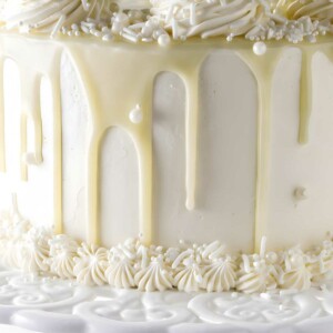 White frosted cake with chocolate drip, swirls, pearls on cake stand.