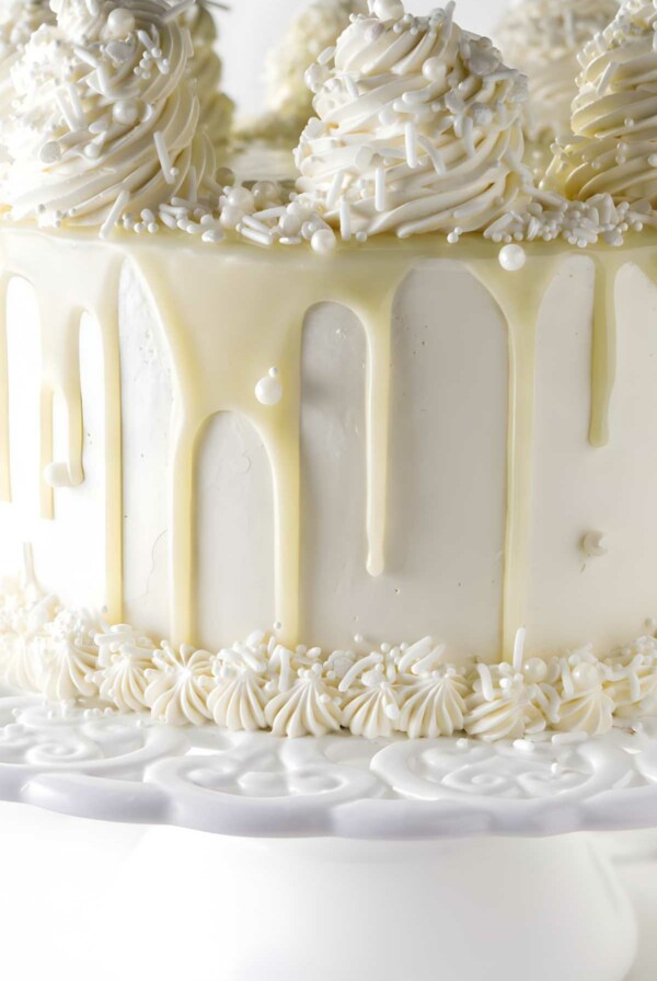 White frosted cake with chocolate drip, swirls, pearls on cake stand.