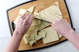Hands chopping white chocolate bars into small pieces on a wooden cutting board.