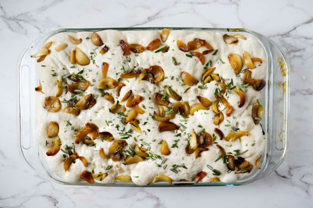 Adding roasted garlic and herbs to a pan of focaccia dough.