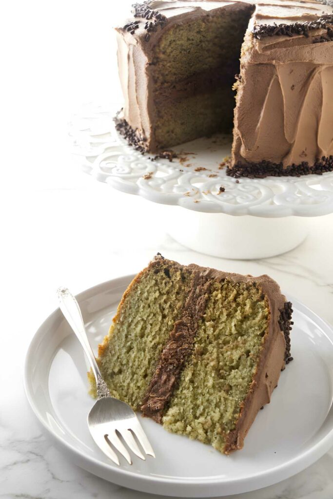 Slice of yellow cake with chocolate frosting on plate; whole cake behind.