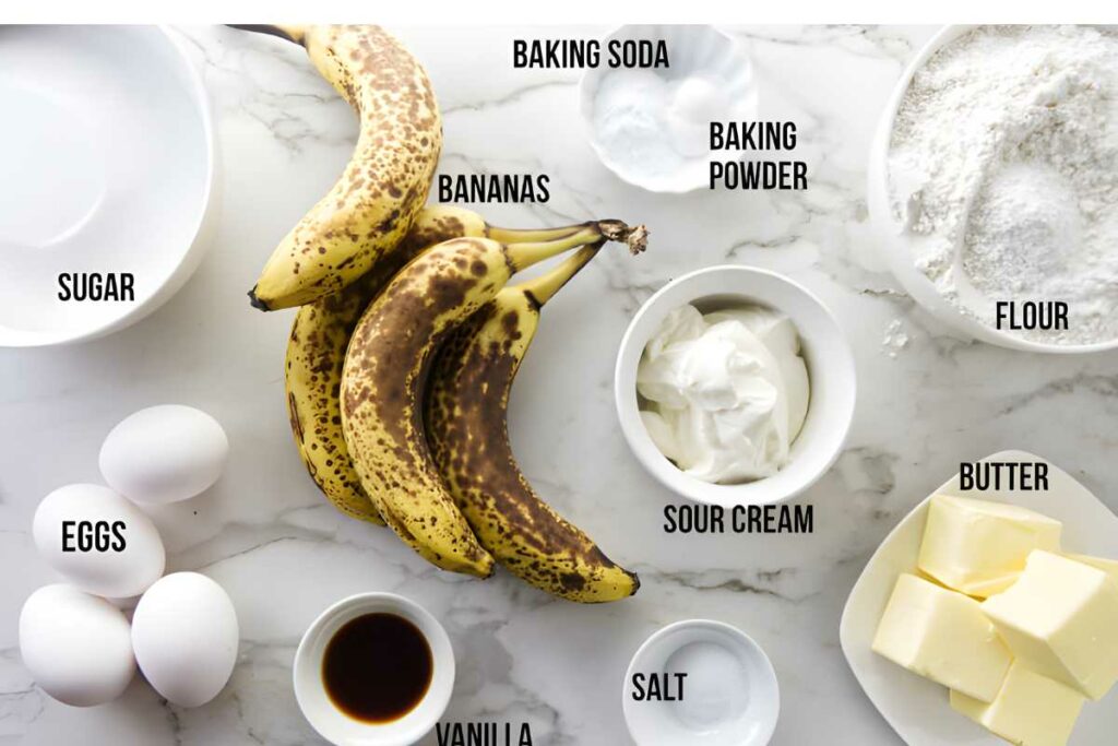 Baking ingredients labeled on marble countertop, including bananas, flour, eggs, and more.