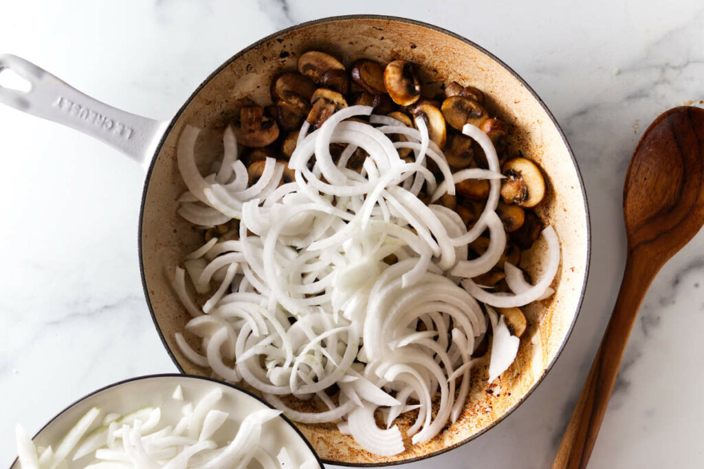 Adding sliced onions to sautéed mushrooms in a skillet as part of the beef stroganoff recipe preparation.