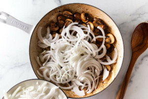 Adding sliced onions to sautéed mushrooms in a skillet as part of the beef stroganoff recipe preparation.