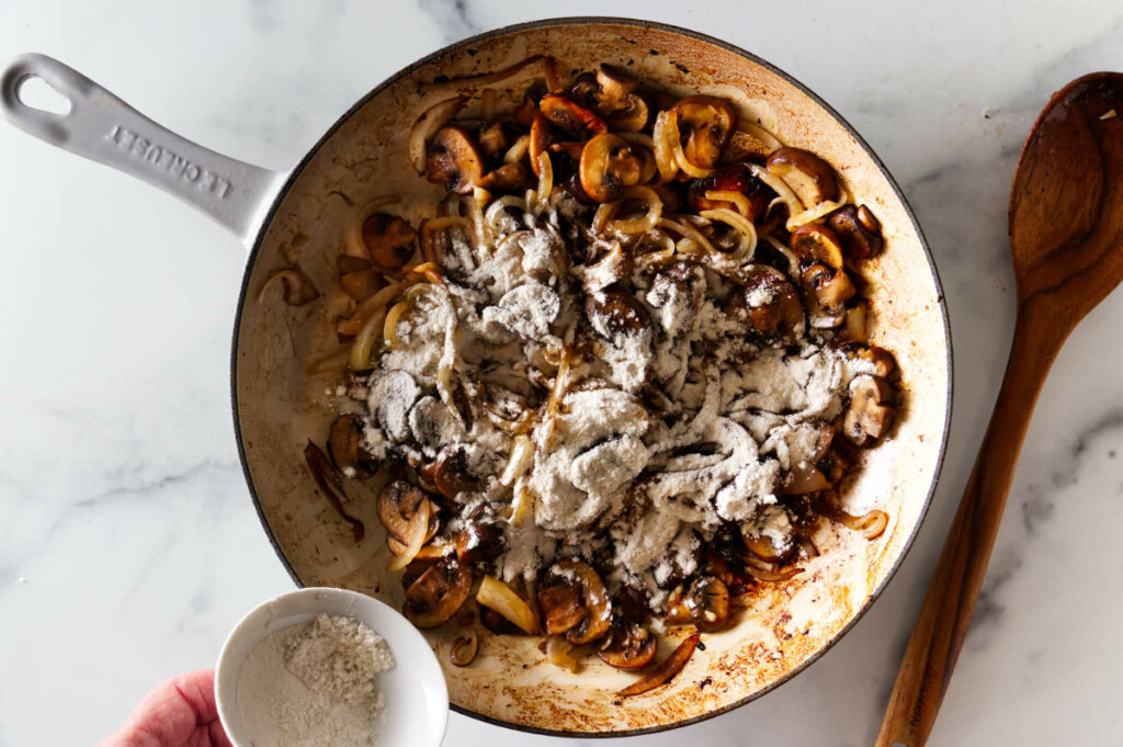 Adding flour to a skillet with mushrooms and onions.