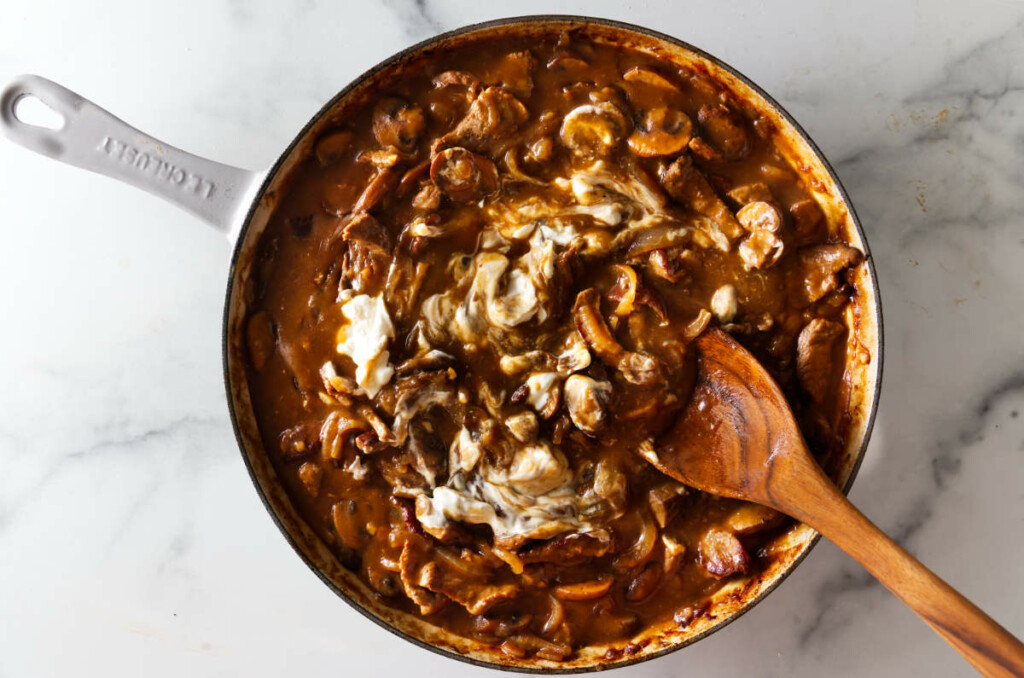 Adding sour cream to the creamy beef, mushroom, and onion mixture to finish the stroganoff sauce.