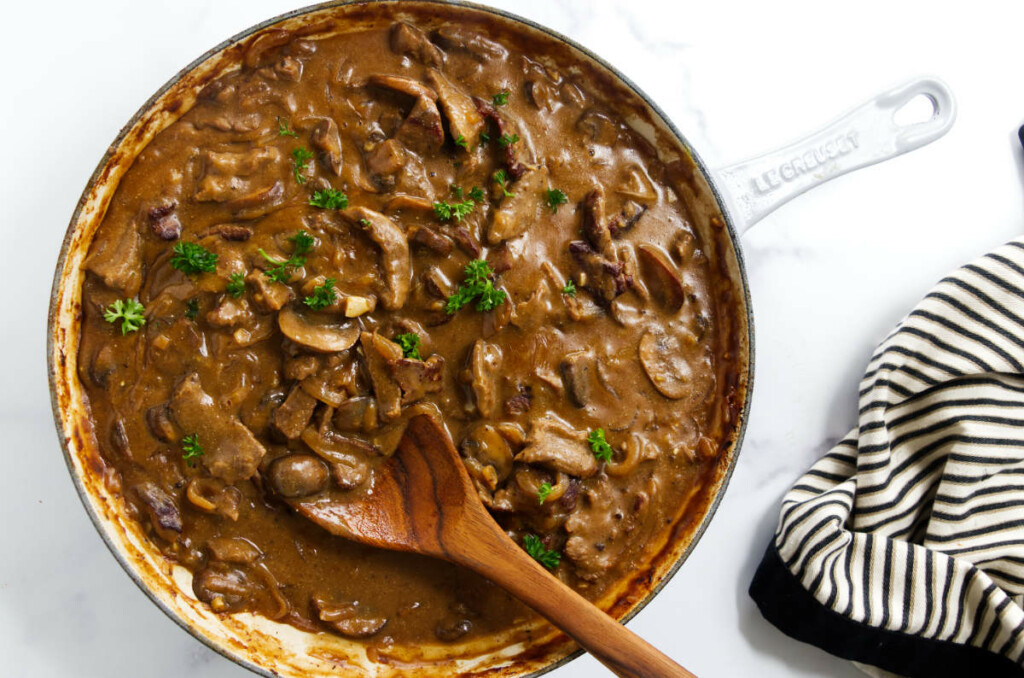 Creamy beef stroganoff sauce with sliced steak, mushrooms, and onions in a skillet.