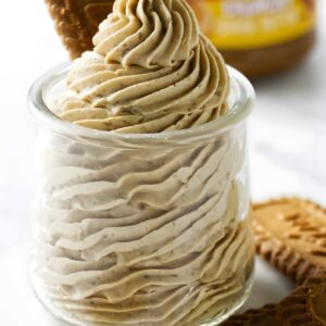 Glass jar of cookie butter frosting topped with Biscoff cookie, cookies nearby.