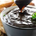 Spoon dripping rich gravy into bowl, parsley garnish, bread blurred behind.