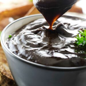 Spoon dripping rich gravy into bowl, parsley garnish, bread blurred behind.