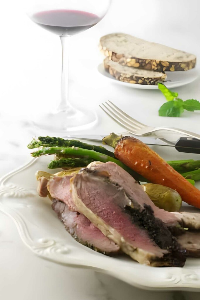 Butterflied leg of lamb with vegetables, bread, and red wine on a white table.