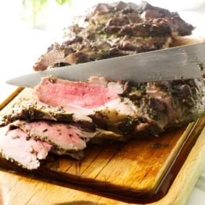 Knife slicing herb-crusted roast lamb, revealing pink interior on wooden board.